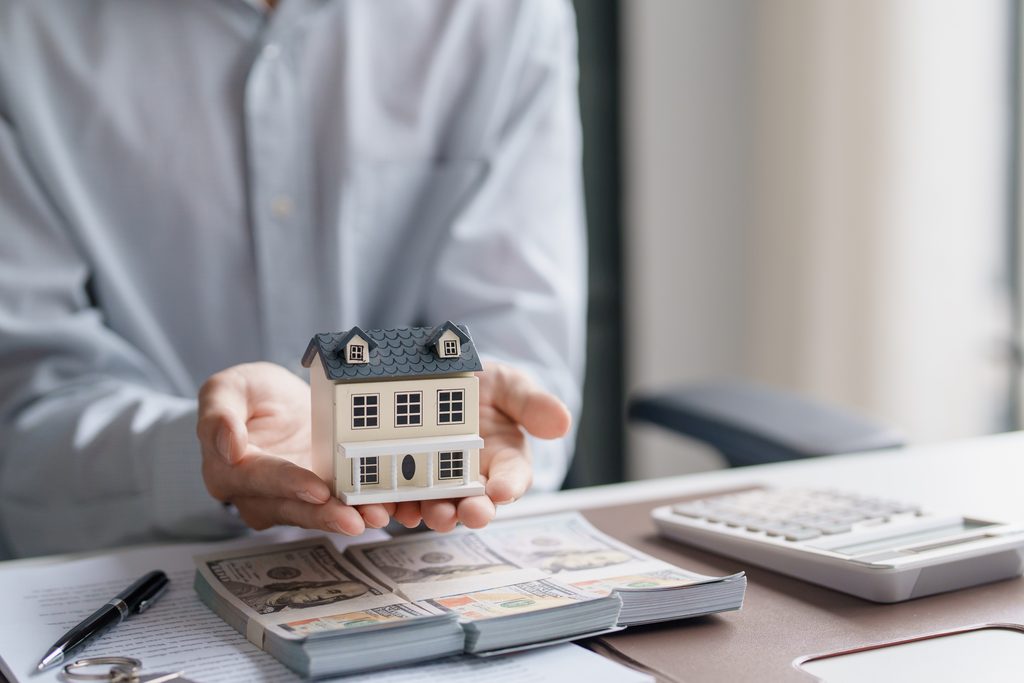 A realtor offering a renter the opportunity to purchase a home, symbolized by a house model, cash, and a calculator.